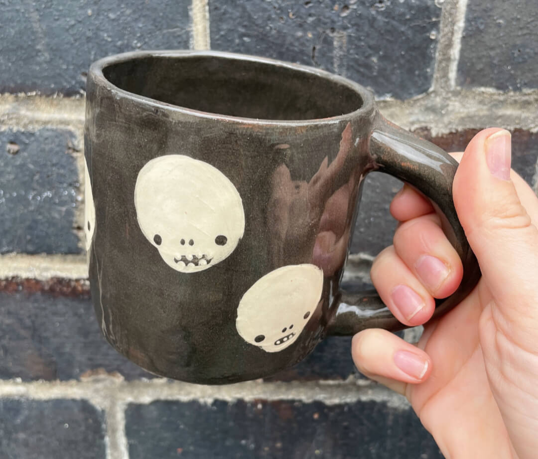 A handmade black terracotta mug with cream skull design by Bristol ceramist Rebecca Edelmann. Held against a dark wall background by a white woman's hand.