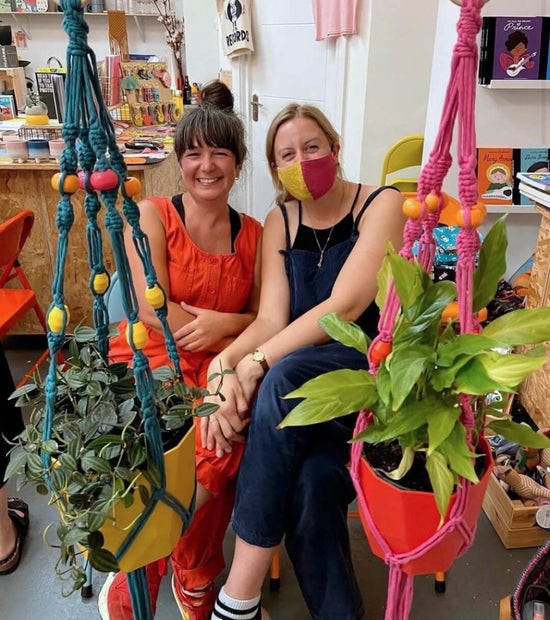 Two white women sit together smiling showing off the macramé plant hangers they have just made in a creative workshop at BAM Store + Space in Bristol