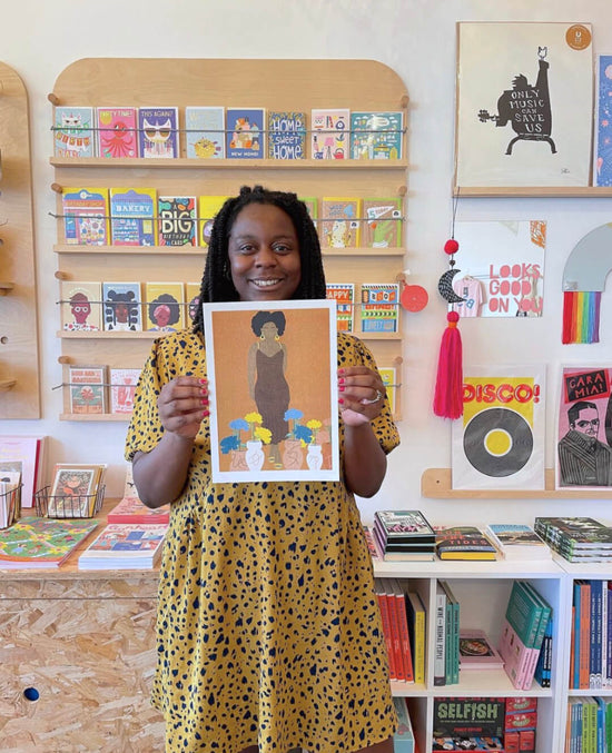 Lucy Turner, a Black artist from Bristol, UK stands holding a limited edition art print against the colourful backdrop of BAM Store + Space, a non-profit independent gift shop in Easton. Lucy is smiling and wearing a yellow dress.