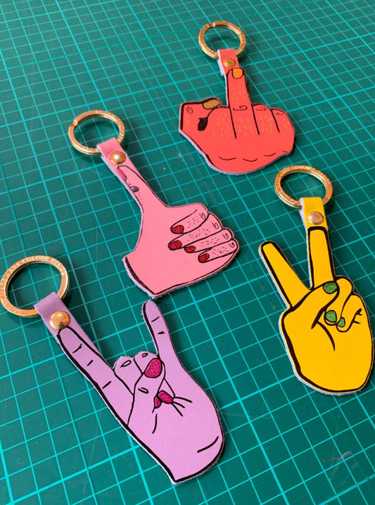 Close up of colourful leather key rings by Ark Colour Design. Featuring hands making a peace sign, a rock on sign, a thumbs up and a cheeky middle finger.
